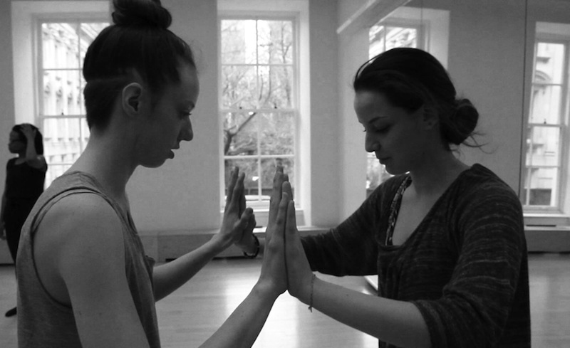 Two dancers face one another and connect their palms 